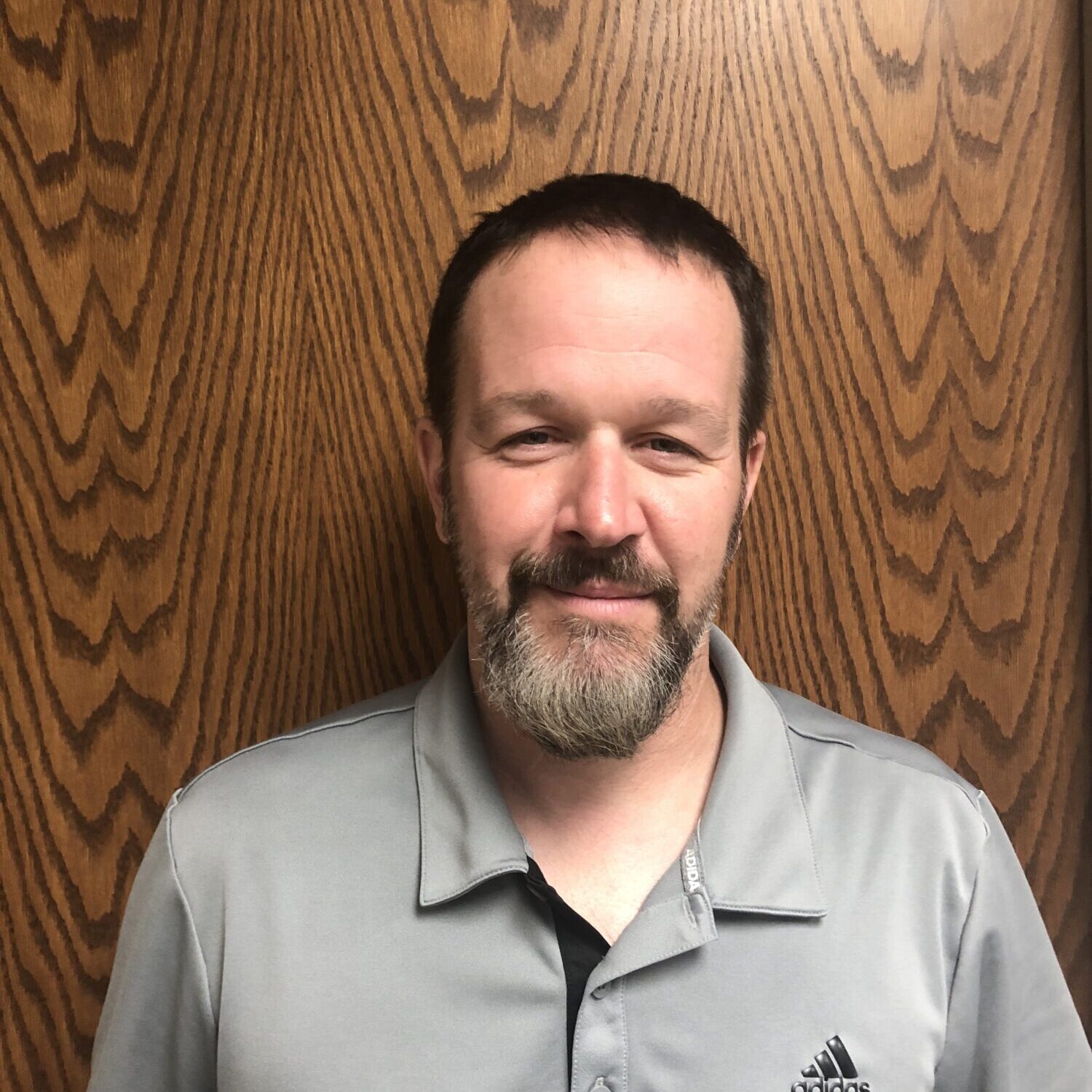 Man smiling in gray shirt with wooden background.